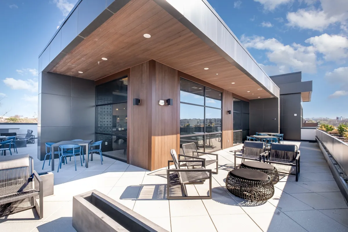 A sophisticated outdoor space featuring modern furniture, large panoramic windows, and a wooden ceiling, with the city skyline visible beyond. The area is ideal for socializing or enjoying a moment of relaxation between work commitments.