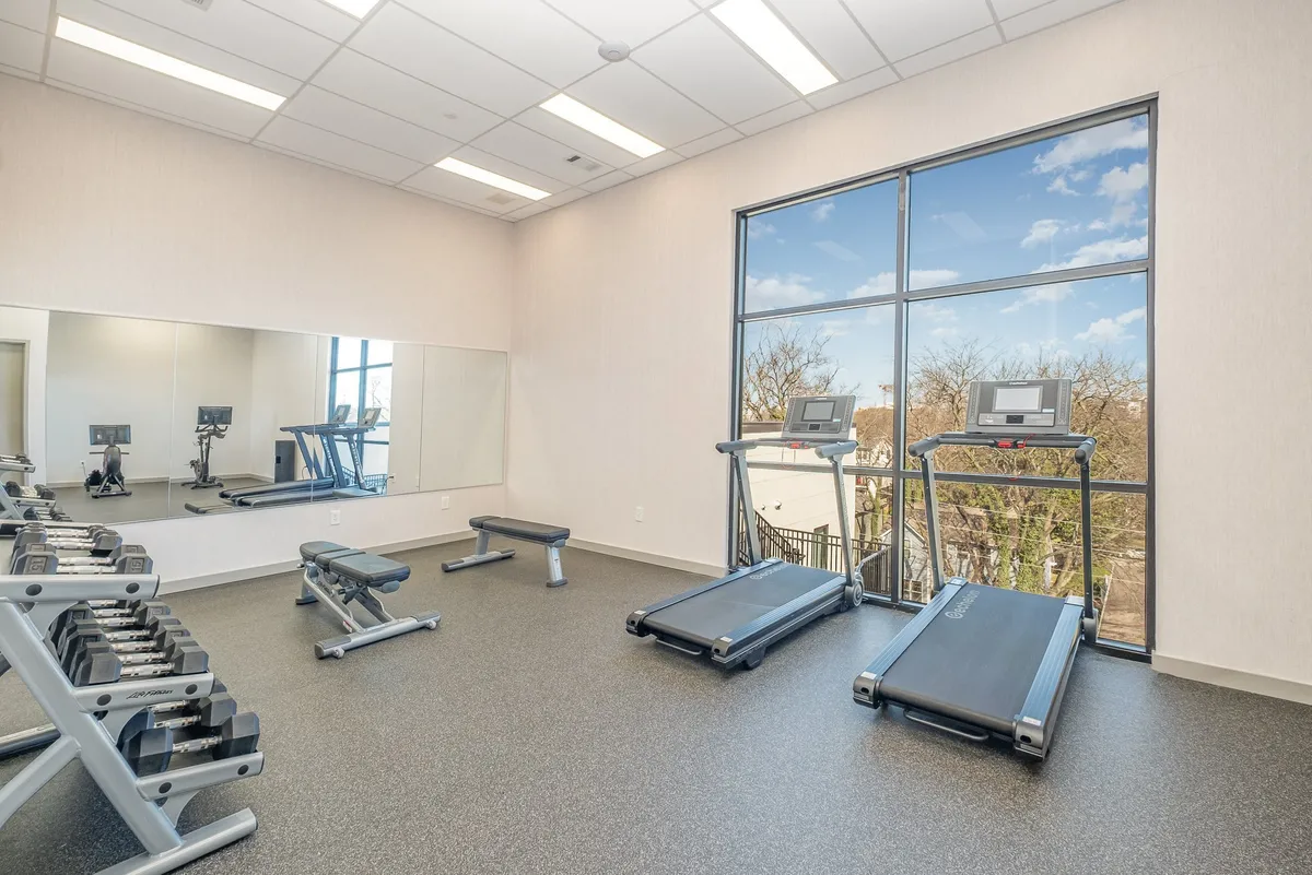 This image depicts a modern, well-lit gym featuring treadmills, weights, large windows, and mirrors. The space is open and inviting, with natural light streaming in and fitness equipment that caters to both cardio and strength training needs.
