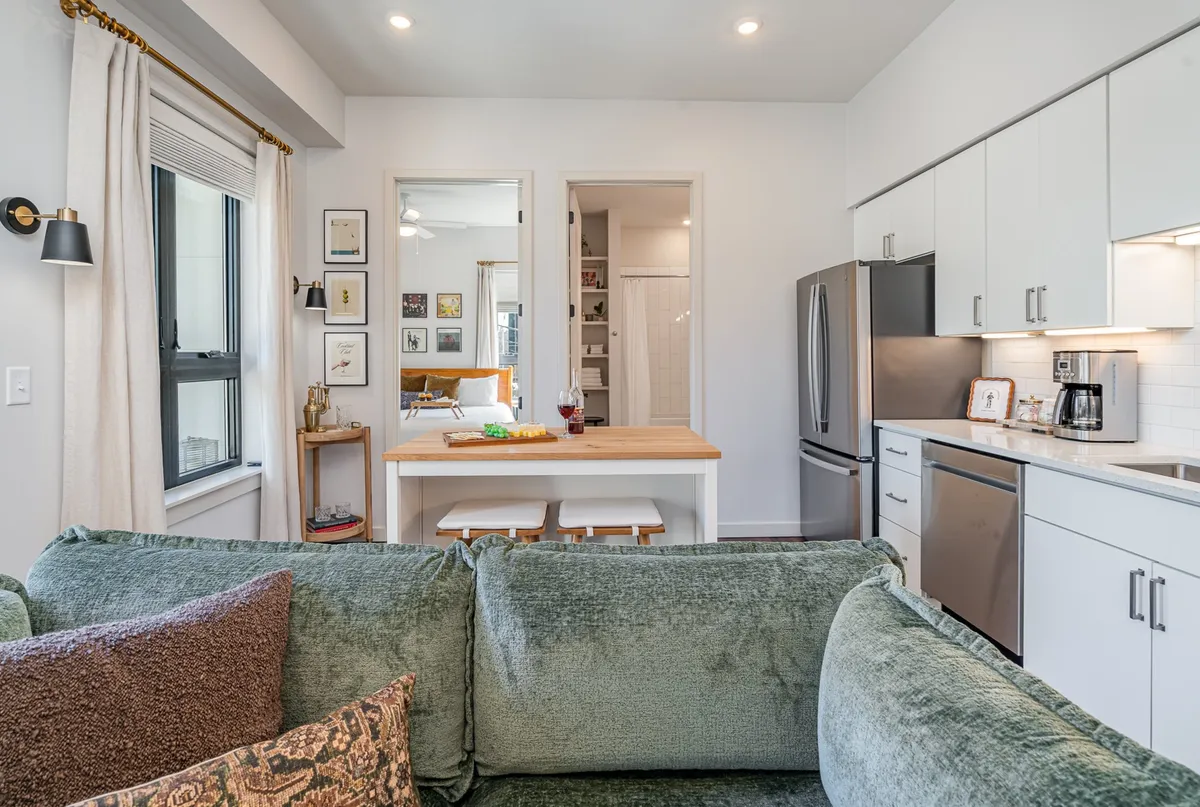 A cozy, sunlit open living area with a comfortable green sofa, a wooden dining table set for meals, and kitchen appliances visible in the background. Framed artwork adds character to the space, enhancing the welcoming, home-like atmosphere.