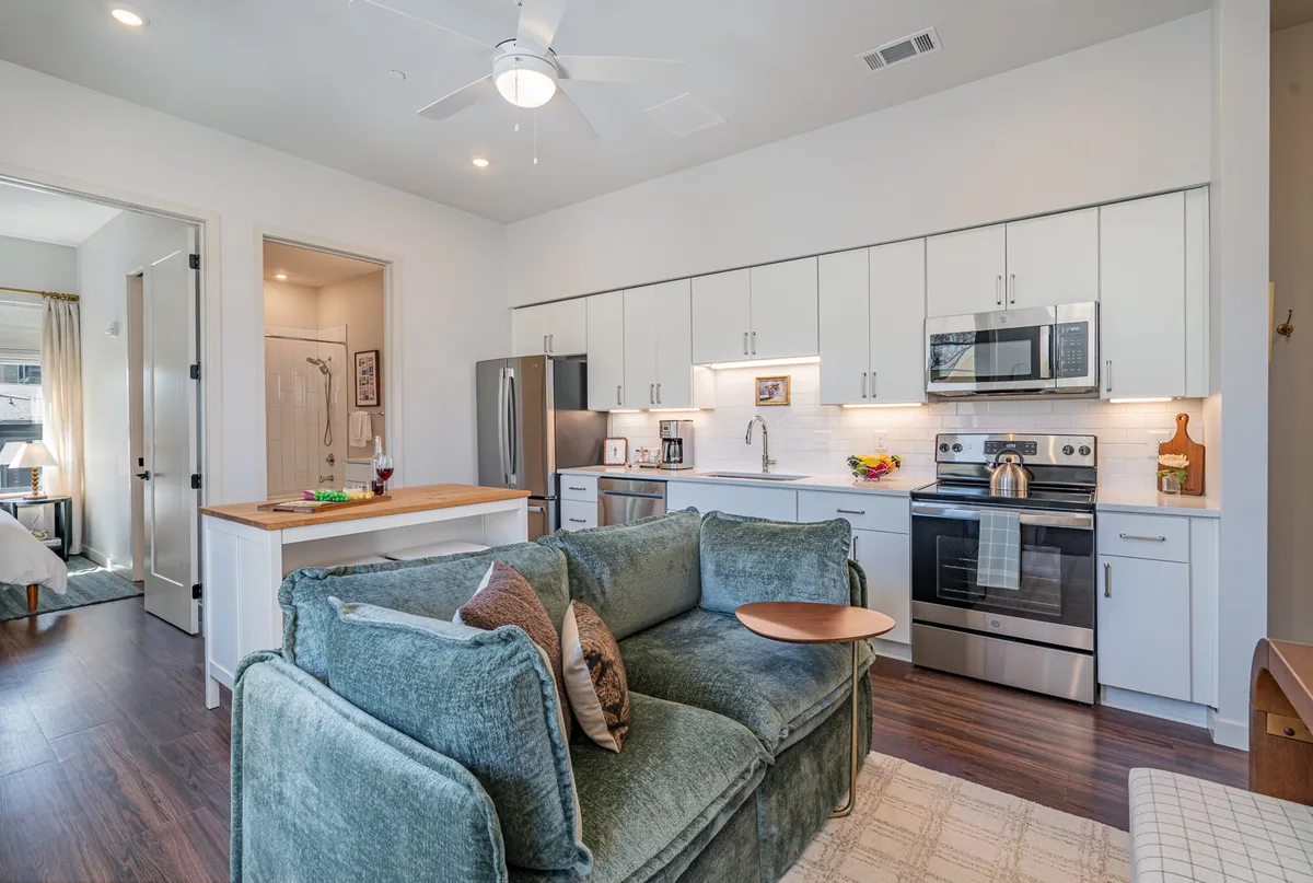A bright and stylish interior scene featuring a green couch facing a kitchen with a central butcher block island and sleek stainless steel appliances. The space is open-concept, with inviting seating ideal for gatherings, and subtle décor notes that reflect a modern, welcoming Nashville vibe.