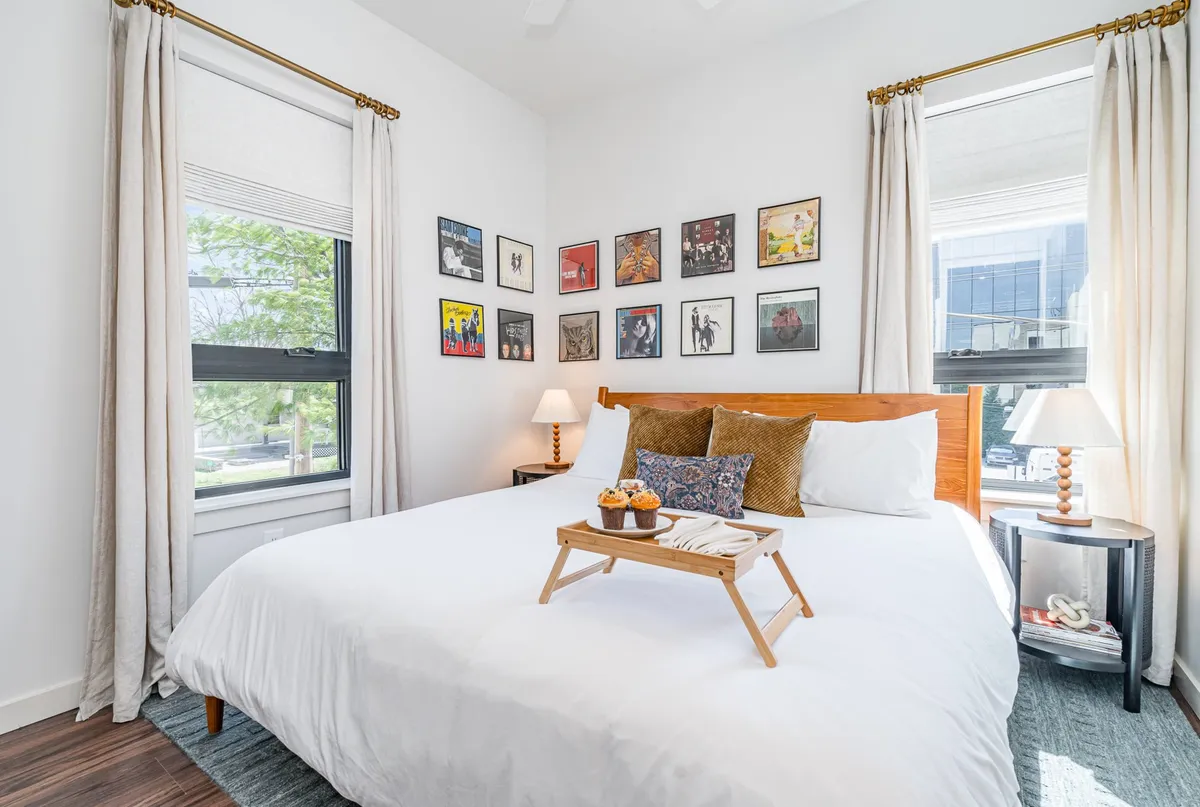 A cozy bedroom featuring a bed with crisp white linens, framed artwork on the walls, wooden bedside tables, decorative pillows, and a tray with snacks, all bathed in gentle natural light.