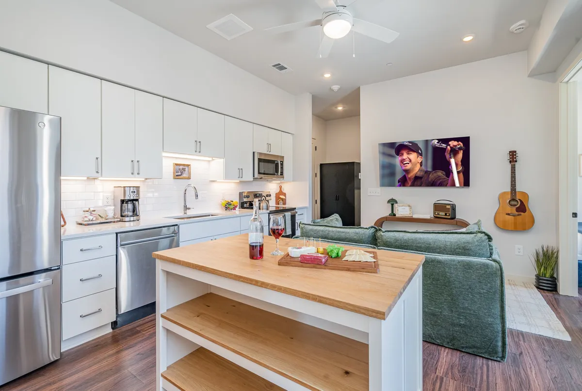 An inviting living and kitchen area scene with a sofa facing a kitchen island adorned with food and drinks, a guitar propped nearby, and a TV displaying a performance. The décor is music-inspired, nodding to Nashville’s Music Row, with a casual and social atmosphere perfect for entertaining or relaxing.