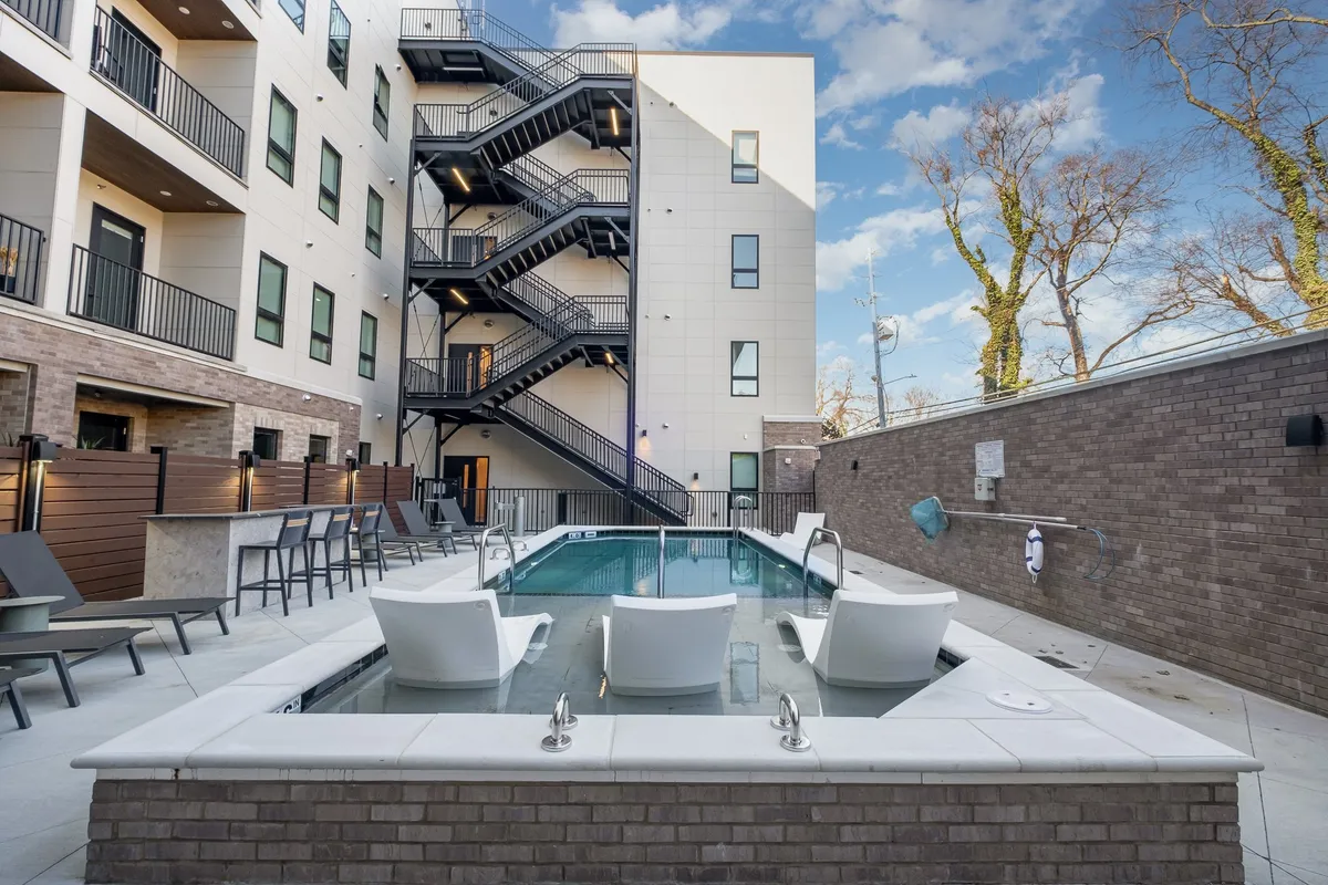 A vibrant exterior shot of a swimming pool surrounded by lounge chairs, an outdoor staircase, and the building facade. The area is bathed in natural light, evoking a lively, resort-style atmosphere perfect for family fun and relaxation.