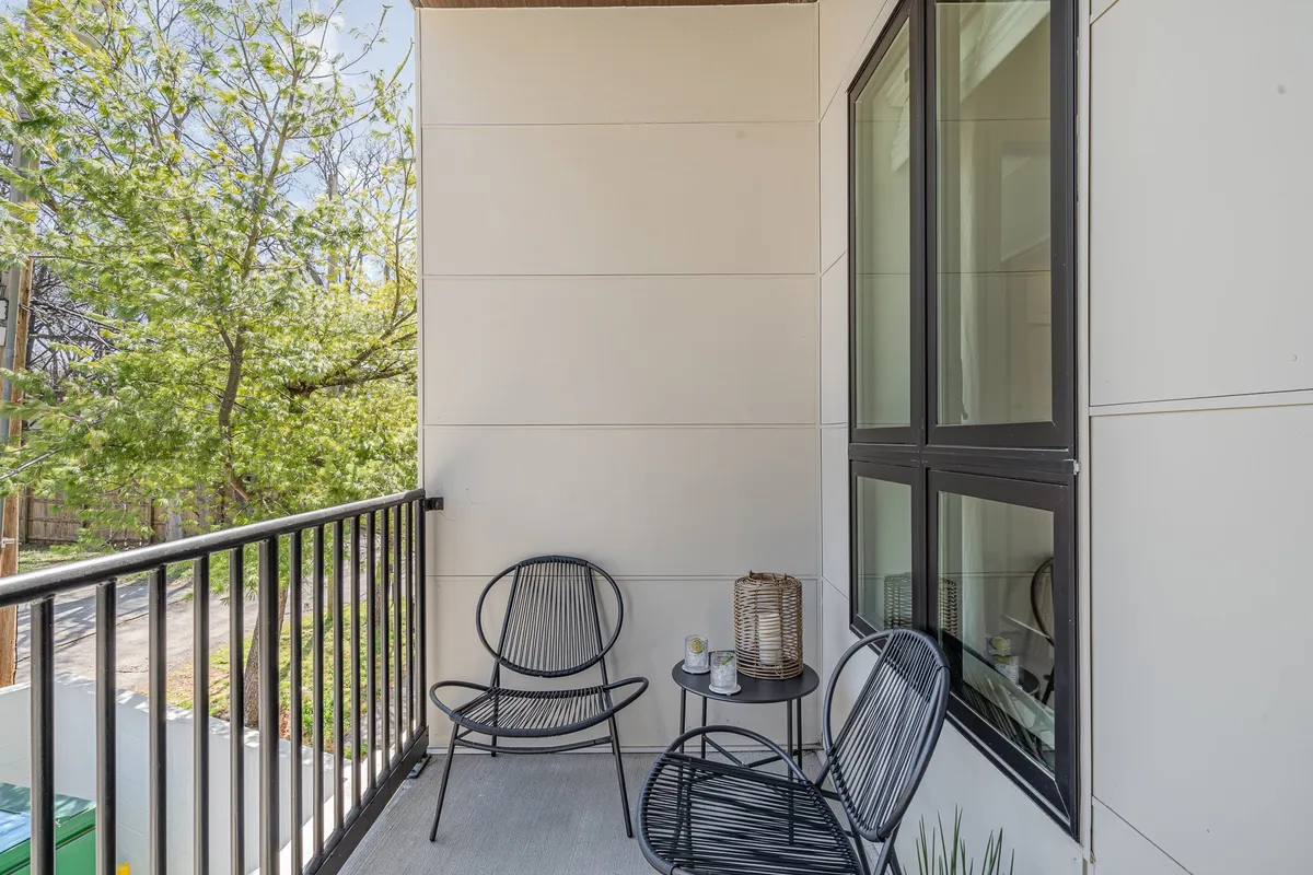 A cozy private balcony space features two comfortable chairs, a small table with a decorative lantern, and lush greenery just beyond the railing. The arrangement is perfect for enjoying morning coffee, reading, or unwinding as daylight fades.