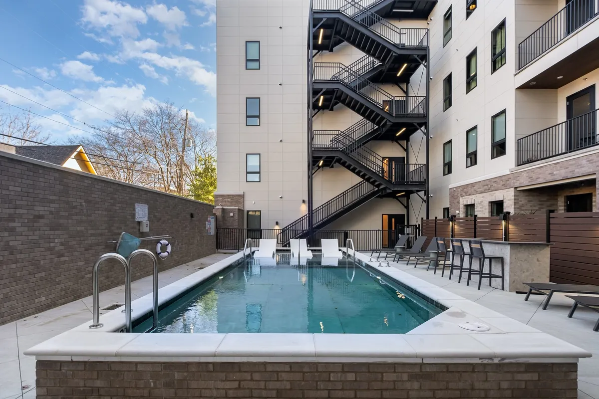 A sun-drenched community swimming pool is framed by a staircase leading down to a deck with lounge chairs, offering a tranquil setting for swimming, sunbathing, and socializing. The inviting water glistens in the light, suggesting both relaxation and recreation.